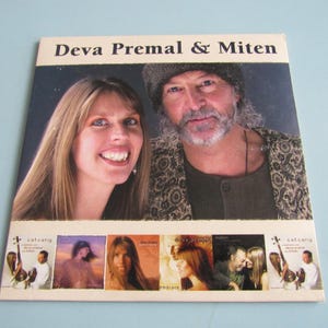 May include: A CD cover featuring the text "Deva Premal & Miten" above a photo of a smiling woman and a man with a beard. Below are smaller images of other album covers. The background is a light blue.