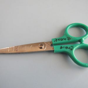 May include: Scissors with gold blades and green handles. The word "LEFTY" is engraved on the blade. The handles have the names "Craig" and "Cheat" written on them in black marker.