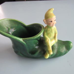 May include: A vintage ceramic planter in the shape of a green shoe, with a seated elf figurine. The elf is wearing a yellow-green outfit and hat. The shoe-shaped planter is a darker shade of green.