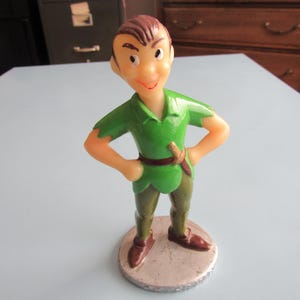 May include: A vintage Peter Pan figurine. The figure is wearing a green tunic, brown pants, and brown shoes. He has brown hair and is standing on a round, silver-colored base. The figurine has a smiling expression and is posed with hands on hips.