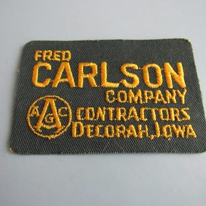 May include: Embroidered patch with the text "FRED CARLSON COMPANY CONTRACTORS DECORAH, IOWA" in gold thread on a dark gray background. The patch has a rectangular shape with rounded corners.
