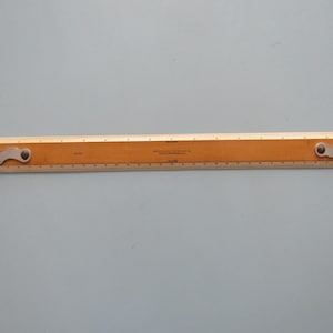 May include: A vintage wooden ruler with metal ends. The ruler is light brown with black markings and measurements in inches. The metal ends are silver and have a decorative shape. The text on the ruler is partially visible.
