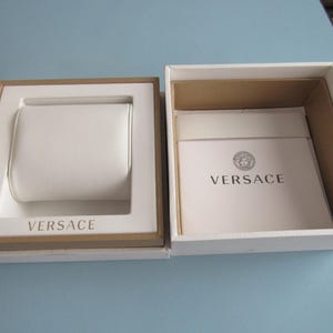 May include: A white and tan square presentation box with a removable lid. The interior features a white watch cushion and a white booklet with the Versace logo and text printed in black. The brand name Versace is also embossed on the white base.