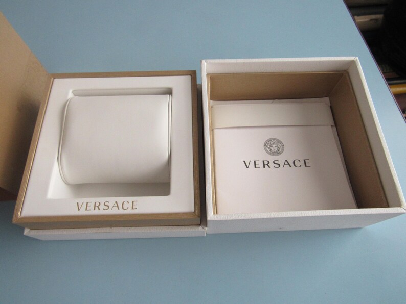 May include: A white and tan square presentation box with a removable lid. The interior features a white watch cushion and a white booklet with the Versace logo and text printed in black. The brand name Versace is also embossed on the white base.
