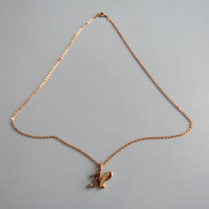May include: A gold-colored necklace with a delicate chain and a pendant shaped like the letter "R". The pendant has a textured surface and is suspended from the chain. The necklace is displayed against a light gray background.