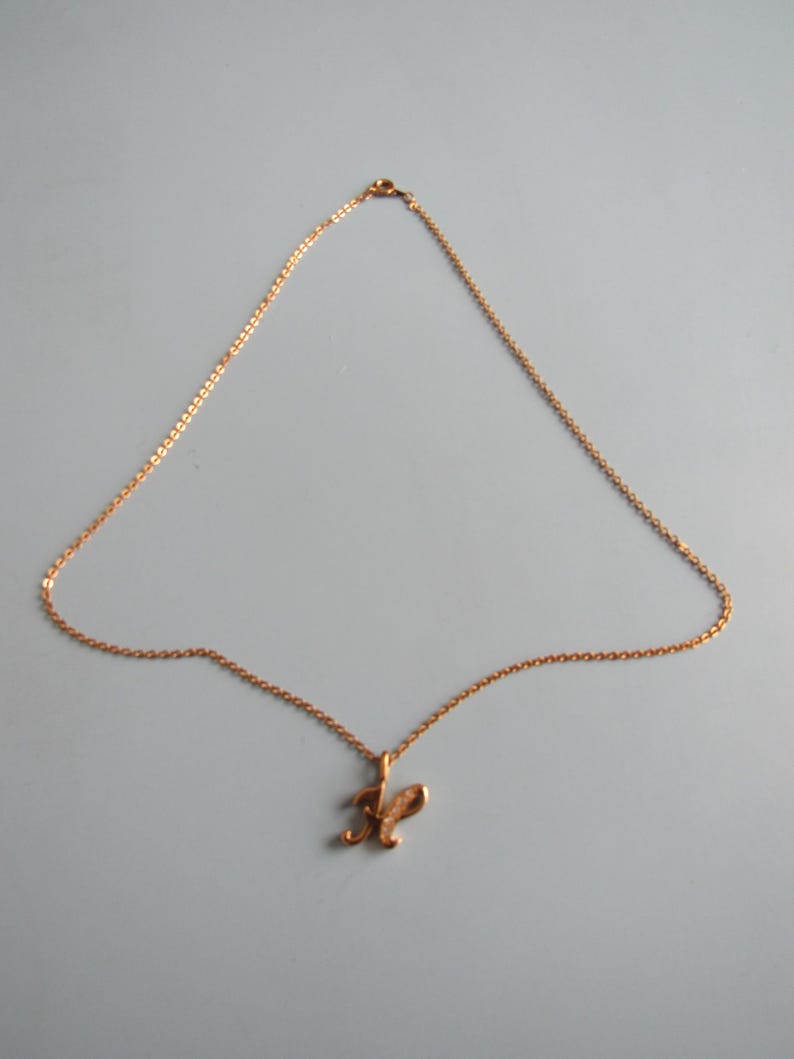 May include: A gold-colored necklace with a delicate chain and a pendant shaped like the letter "R". The pendant has a textured surface and is suspended from the chain. The necklace is displayed against a light gray background.