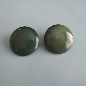 May include: A pair of circular, domed buttons featuring a glossy, mottled finish in shades of dark forest green and muted olive. The surface of each button is smooth and reflective, showcasing subtle variations in color depth and tone throughout.