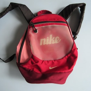 May include: A red and pink Nike backpack featuring a rounded shape, a top handle, and dark brown adjustable shoulder straps. The front displays the word nike in white lettering, a small white swoosh logo, and a zippered compartment with a mesh overlay.