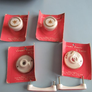 May include: A collection of vintage ceramic cabinet hardware, including knobs and pulls. The knobs are white with a gold starburst design. The pulls and rosettes are also white with gold accents. Packaged on red and white striped cards.