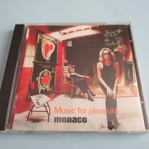 May include: A compact disc jewel case featuring the album Music for Pleasure by the band Monaco. The cover art displays a red-toned interior scene with two figures, one standing in the foreground and one seated on a chair in the background.