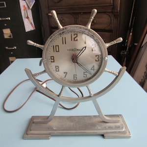 May include: A silver-colored electric clock with a ship's wheel design. The clock face has black numbers and hands, and the words "Electric Pennant" are printed on the face. The clock is on a silver-colored stand with a curved base.
