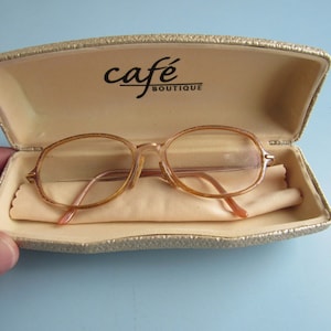 May include: A beige eyeglasses case with a brown pair of glasses inside. The case has the text "cafe BOUTIQUE" printed on the top.