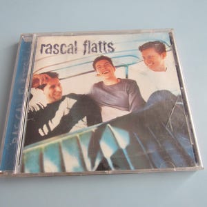 May include: A CD case with a photo of three men in a car. The case is blue and silver and has the text "Rascal Flatts" on the front.