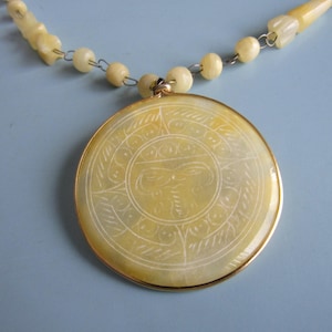 May include: A round, pale yellow pendant necklace featuring an intricate, etched circular design. The pendant is encased in a thin gold-toned metal rim and is suspended from a chain composed of matching yellow beads and small metal links.