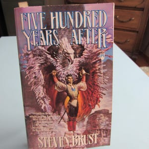 May include: A paperback book titled Five Hundred Years After by Steven Brust. The cover features a central figure with arms raised, standing before a large, feathered creature with wings spread. The background shows a rocky landscape in shades of purple.