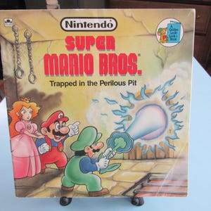 May include: A vintage Super Mario Bros. book titled "Trapped in the Perilous Pit." The cover features Mario, Luigi, and Princess Peach in a colorful illustration. The Nintendo logo is at the top, with a "Golden Look-Look Book" emblem.