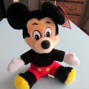May include: A plush Mickey Mouse toy, featuring black ears, a black shirt, red shorts, and yellow shoes. The toy has a beige face, white gloves, and a black nose. A red tag is attached to the toy.