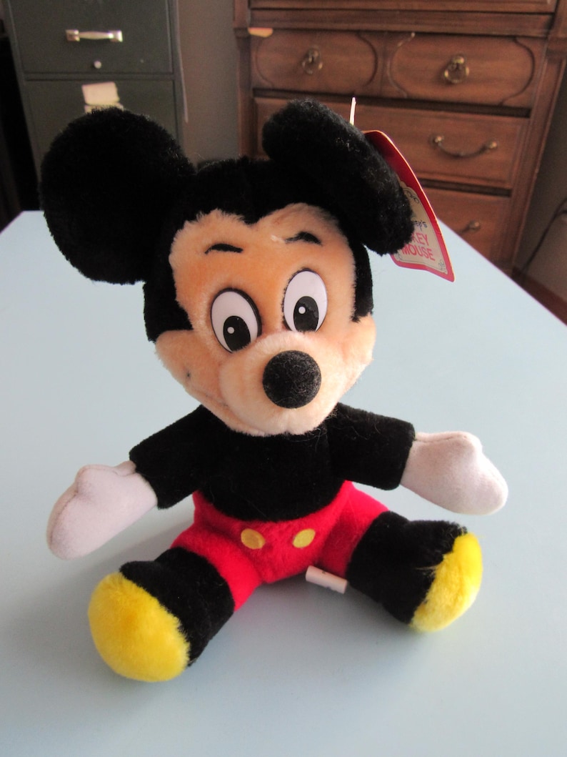 May include: A plush Mickey Mouse toy, featuring black ears, a black shirt, red shorts, and yellow shoes. The toy has a beige face, white gloves, and a black nose. A red tag is attached to the toy.