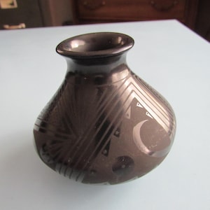 May include: A dark brown ceramic vase with a rounded body and a narrow neck. The vase features etched geometric patterns and a crescent moon design. The vase is likely handmade and suitable for home decor.