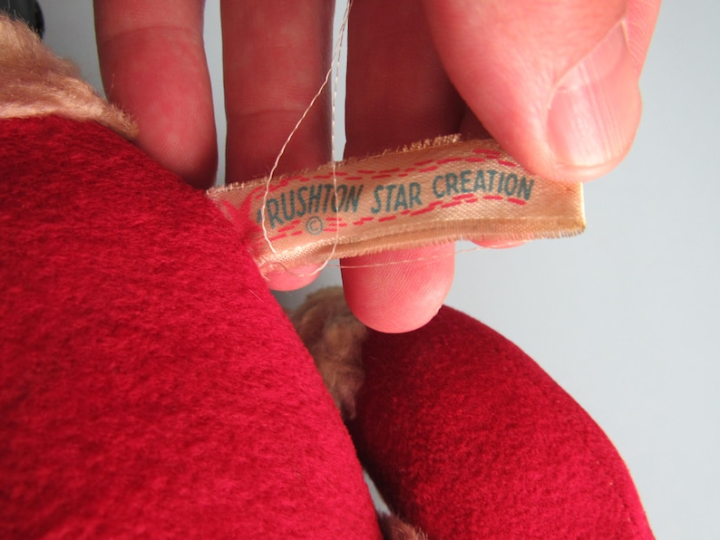 May include: Close-up of a plush toy with a red felt body and a tag that reads "RUSHTON STAR CREATION" in green text. The tag is attached with a white thread. The toy has a beige-colored area.