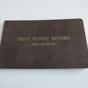 May include: A brown Pilot Flight Record and Log Book. The cover is a dark brown color with gold lettering. The text on the cover reads "PILOT FLIGHT RECORD AND LOG BOOK". The book is rectangular in shape.