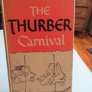 May include: A vintage hardcover book titled "The Thurber Carnival." The cover features a red background with black and gold lettering. Below the title is a tan illustration of carousel figures. The book has a red spine.