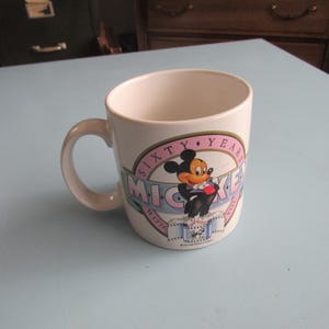 May include: A white ceramic mug featuring a graphic of Mickey Mouse in a tuxedo. The design includes a pink circular border with the text SIXTY YEARS and a blue banner with the text MICKEY WITH YOU, along with a film strip illustration at the base.