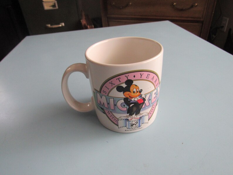 May include: A white ceramic mug featuring a graphic of Mickey Mouse in a tuxedo. The design includes a pink circular border with the text SIXTY YEARS and a blue banner with the text MICKEY WITH YOU, along with a film strip illustration at the base.