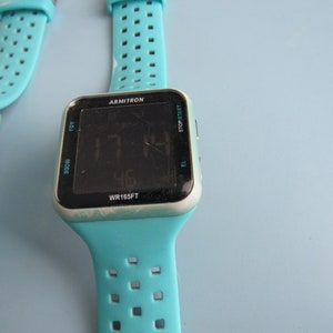 May include: Two Armitron digital watches with turquoise bands. The watch faces display the time and have buttons for adjusting settings. The watch cases are silver-toned. The watch bands have a series of small holes for breathability.