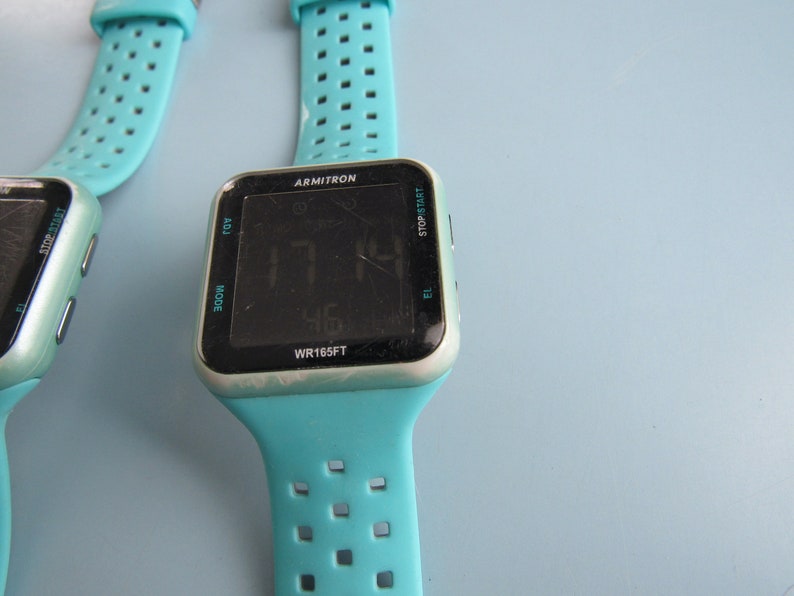 May include: Two Armitron digital watches with turquoise bands. The watch faces display the time and have buttons for adjusting settings. The watch cases are silver-toned. The watch bands have a series of small holes for breathability.