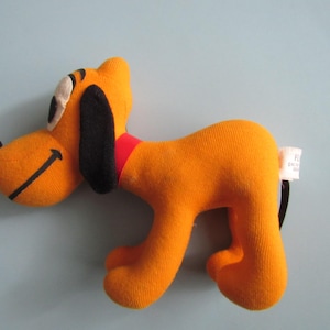 May include: Orange plush dog toy with black ears, a black nose, and a red collar. The dog has a black tail and a tag attached to its rear. The toy is a cartoon character.