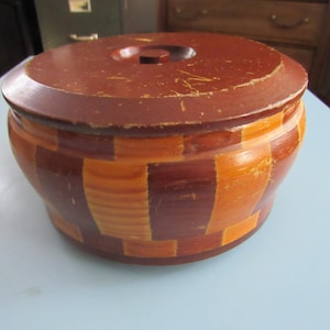 May include: A round wooden container with a lid. The container is made of alternating dark brown and orange-brown rectangular blocks. The lid is dark brown with a small, round handle. The container sits on a light blue surface.