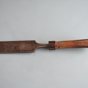 May include: A wood chisel featuring a wide, flat, weathered metal blade with a dark patina. The tool includes a cylindrical wooden handle with a worn finish and a dark metal ferrule connecting the blade to the handle, showing signs of age.