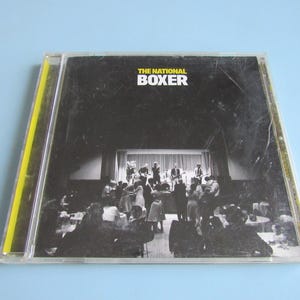 May include: A clear plastic CD jewel case containing the album Boxer by The National. The cover art features a black and white photograph of a band performing on a stage in front of a dancing crowd, with the text THE NATIONAL BOXER in white and yellow.