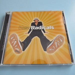 May include: A CD case featuring the album cover for New Radicals. The cover art shows a person in a black outfit and hat, with their legs spread wide. The background is a yellow and orange burst design. The text "New Radicals" is displayed.
