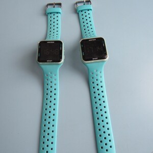 May include: Two digital wristwatches with light blue straps and black square faces. The watch displays the time. The straps have a series of small square perforations. The watch face has the brand name "Armitron" printed on it.