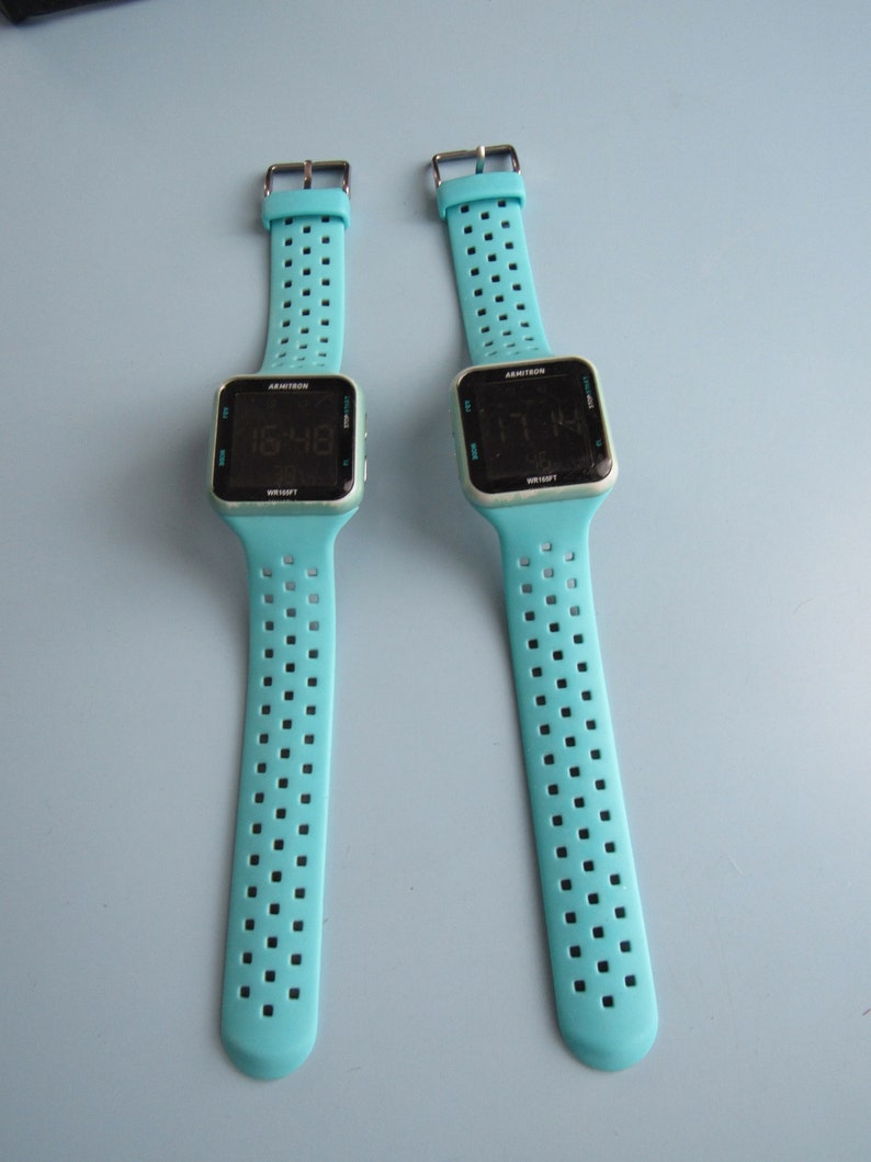 May include: Two digital wristwatches with light blue straps and black square faces. The watch displays the time. The straps have a series of small square perforations. The watch face has the brand name "Armitron" printed on it.