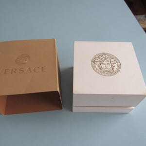 May include: A tan cardboard sleeve with the word VERSACE and a Medusa head logo embossed on the top, positioned next to a white square box featuring a gold Medusa head emblem centered on the lid. Both items are rigid containers for storage.