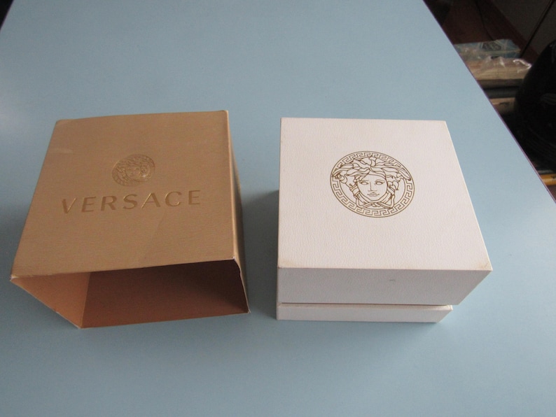 May include: A tan cardboard sleeve with the word VERSACE and a Medusa head logo embossed on the top, positioned next to a white square box featuring a gold Medusa head emblem centered on the lid. Both items are rigid containers for storage.