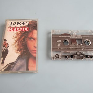 May include: An INXS Kick cassette tape with its clear plastic case. The cover features the band name and album title in red and black text over a portrait of a person with long hair. The cassette itself is clear with a grey label and text.