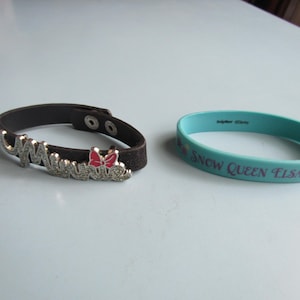 K&ouml;nnte beinhalten: Zwei Armb&auml;nder. Eines ist eine dunkelbraune Leder-Manschette mit dem Namen "Minnie" in silbernen Buchstaben und einer roten Schleife. Das andere ist ein hellblaues Gummiarmband mit dem Text "Snow Queen Elsa".