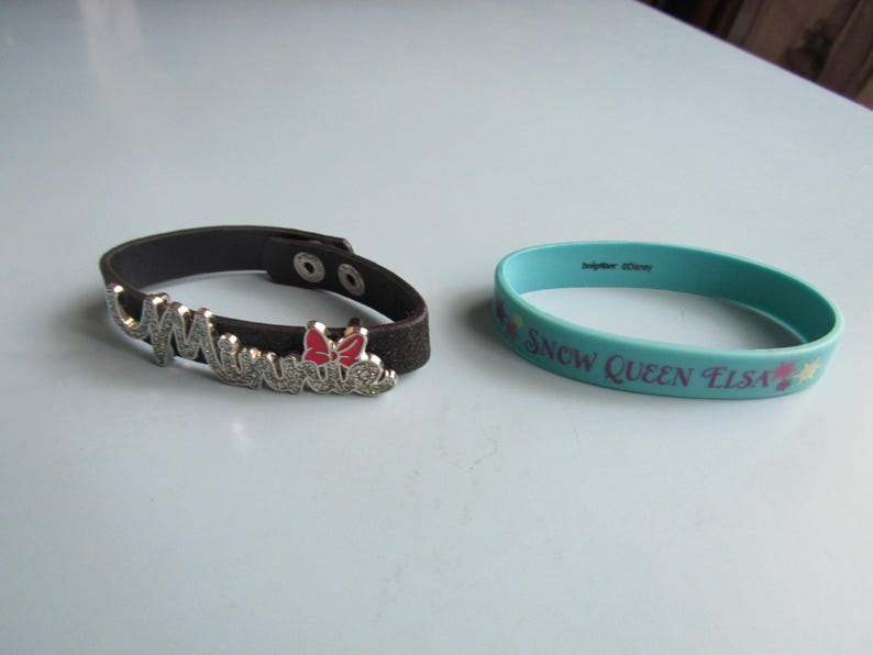 K&ouml;nnte beinhalten: Zwei Armb&auml;nder. Eines ist eine dunkelbraune Leder-Manschette mit dem Namen "Minnie" in silbernen Buchstaben und einer roten Schleife. Das andere ist ein hellblaues Gummiarmband mit dem Text "Snow Queen Elsa".