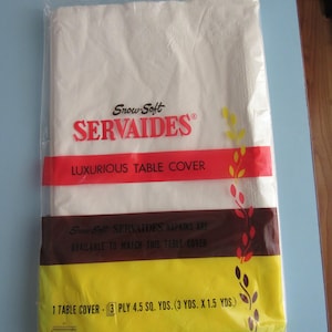 May include: A white plastic-wrapped package containing a white table cover. The package is labeled "Snow-Soft SERVAIDES LUXURIOUS TABLE COVER". The package also states that "SERVAIDES NAPKINS ARE AVAILABLE TO MATCH THIS TABLE COVER" and that the table cover is "1 TABLE COVER - 3 PLY 4.5 SQ. YDS. (3 YDS. X 1.5 YDS.)". The package is labeled with the "ERVING PAPER MILLS" logo.