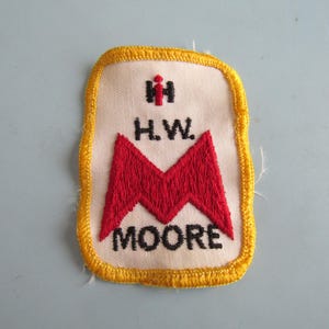 May include: A rectangular embroidered patch with a white background and a yellow border. It features a red stylized letter M, the black text H.W. MOORE, and a small red and black logo at the top. The embroidery is dense and textured throughout.