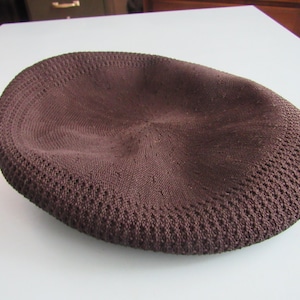 May include: A brown knitted cap with a rounded crown and a textured, perforated brim. The cap is made of a soft, woven material and is designed to fit snugly on the head. The cap is a solid colour and appears to be new.