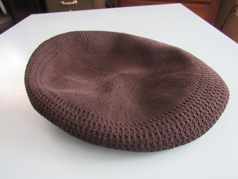May include: A brown knitted cap with a rounded crown and a textured, perforated brim. The cap is made of a soft, woven material and is designed to fit snugly on the head. The cap is a solid colour and appears to be new.