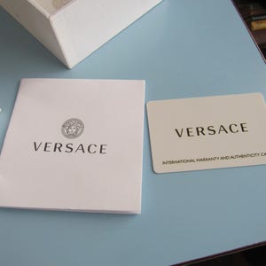 May include: A white Versace booklet with the Medusa head logo and the brand name printed in black, alongside a rectangular off-white Versace international warranty and authenticity card with black text, positioned near a white box corner.
