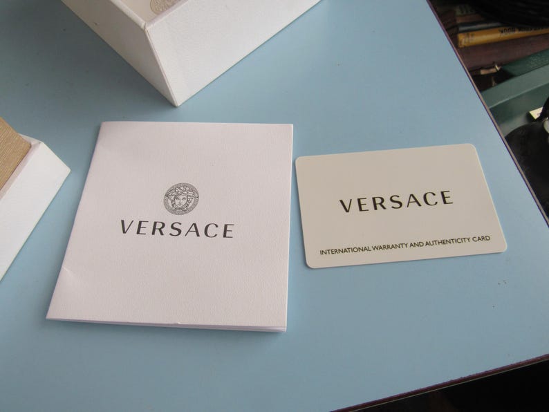 May include: A white Versace booklet with the Medusa head logo and the brand name printed in black, alongside a rectangular off-white Versace international warranty and authenticity card with black text, positioned near a white box corner.