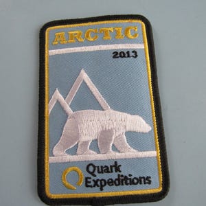May include: A blue and black embroidered patch with the text "Arctic 2013" and a white polar bear walking on a white mountain range. The text "Quark Expeditions" is below the bear with a stylized "Q" in front of the text.