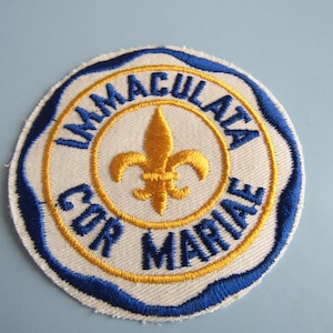 May include: Embroidered patch with a circular design. The patch features a white background with blue and gold stitching. The text "IMMACULATA COR MARIAE" is embroidered in blue. A gold fleur-de-lis is in the center.